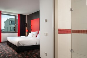 Camera d'albergo con un letto, una parete decorata in rosso, una finestra con tende e un box doccia piastrellato.