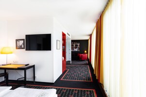 Quarto de hotel com cama, escrivaninha, televisão e uma cadeira vermelha. Há um corredor com carpete e uma porta vermelha que dá para outro quarto.