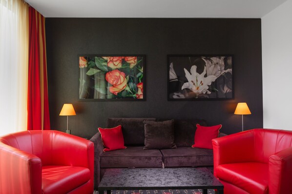 Una sala moderna con un sofá de color oscuro, dos sillones rojos y dos lámparas fijadas a la pared. Encima del sofá, hay dos cuadros con motivos florales.