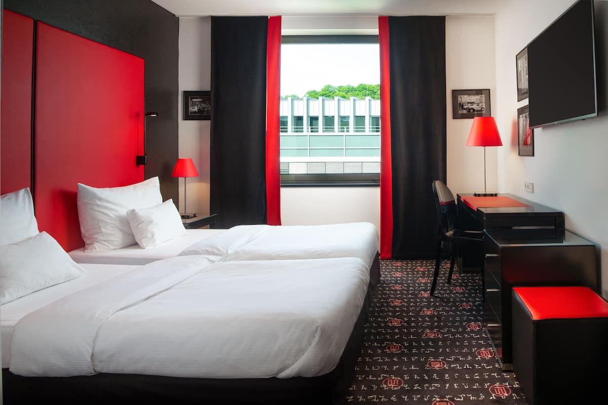 Une chambre d’hôtel avec deux lits, un bureau, une chaise et une fenêtre donnant sur des immeubles et des espaces verts.