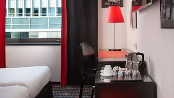Quarto de hotel com cama, escrivaninha com abajur vermelho, cafeteira e janela com vista para edifícios.