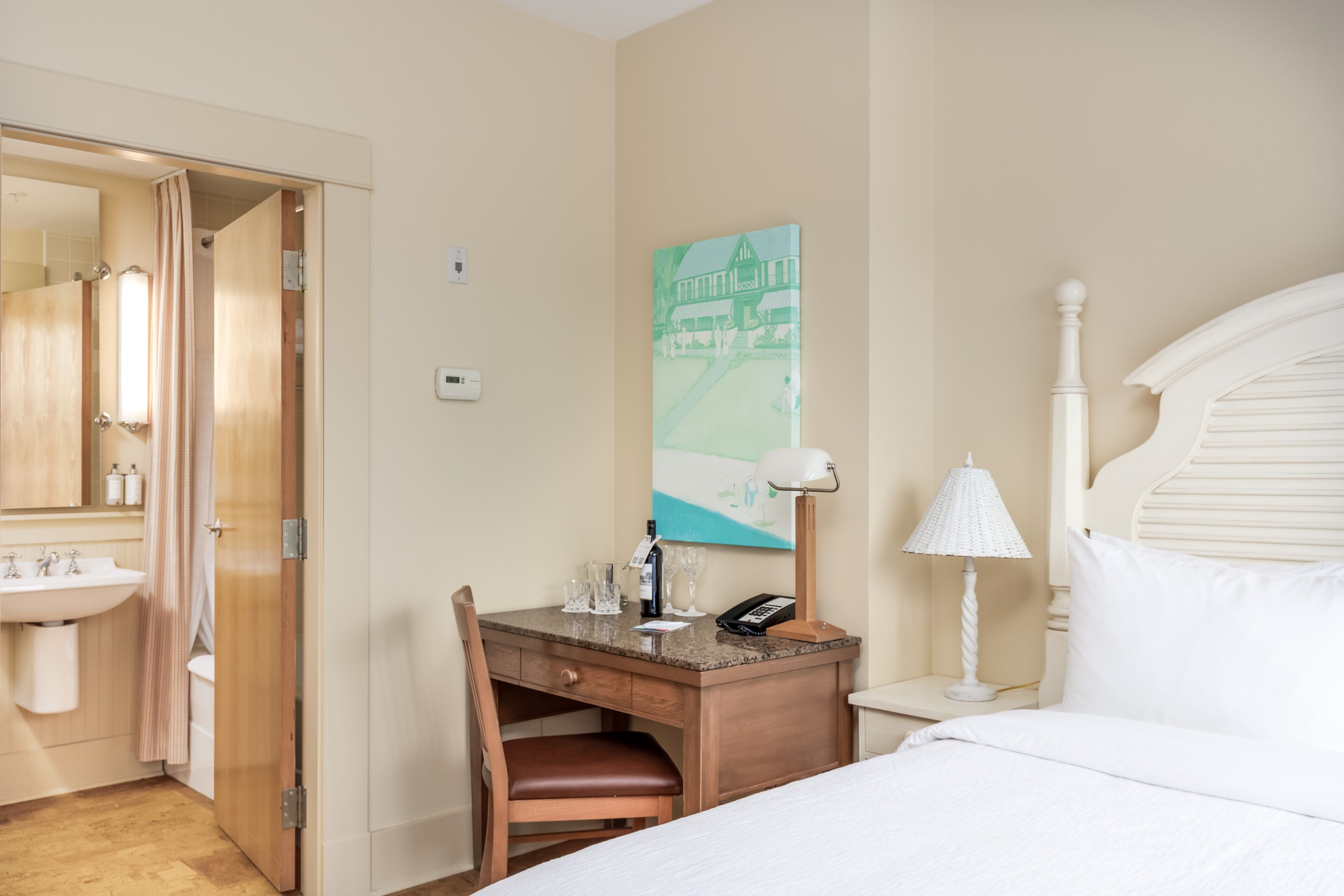 A hotel room with a wooden desk, a bed, a nightstand, a lamp, a painting, a bathroom with a sink and mirror, and a door.