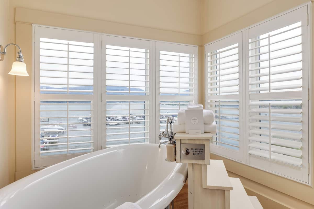 Ein Badezimmer mit einer großen Badewanne, weißen Armaturen und Blick auf den Yachthafen durch große Fenster mit Jalousien.