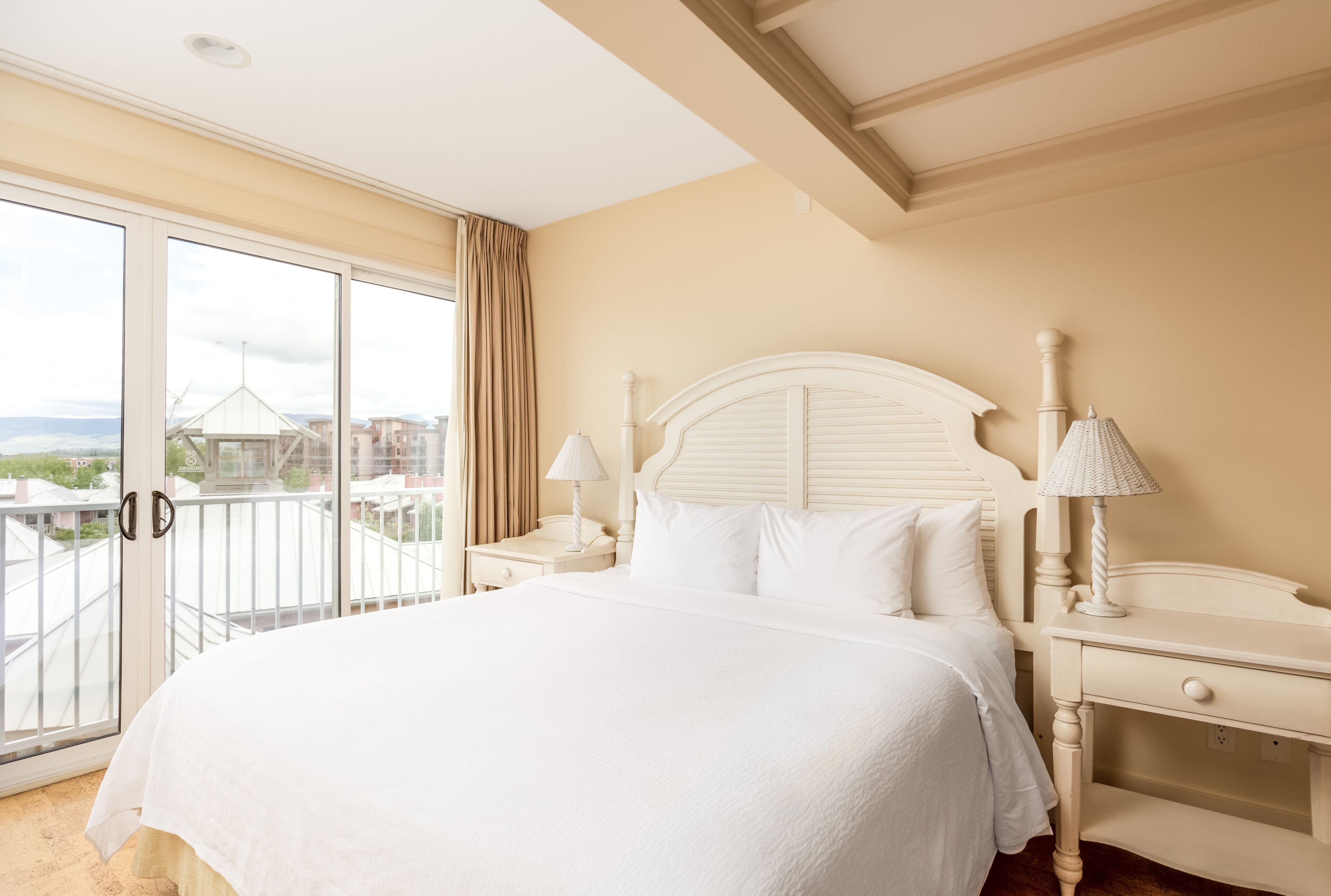 A hotel room with a large bed, two bedside tables, a window with a view of buildings, and a balcony.