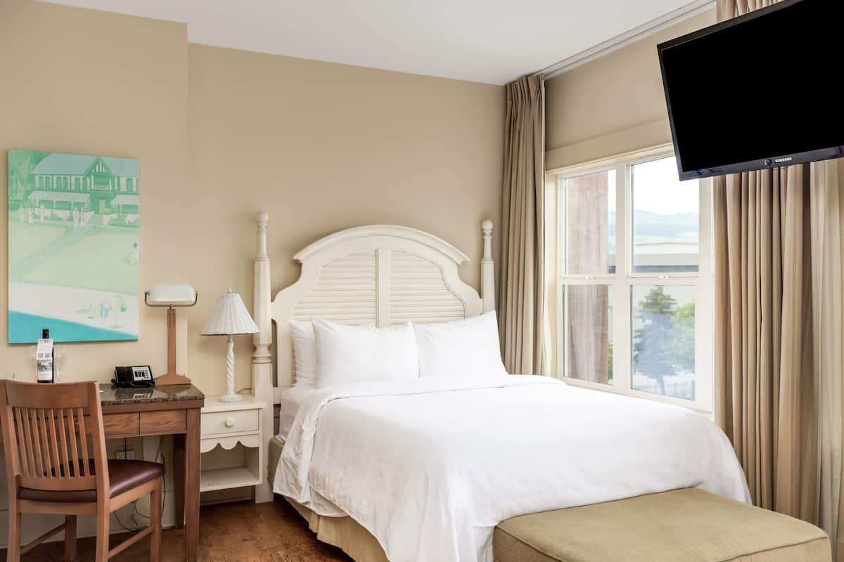 Ein Hotelzimmer mit einem Bett, einem Schreibtisch mit Stuhl, einer Lampe, einem Gemälde an der Wand und einem an der Wand befestigten Fernseher.