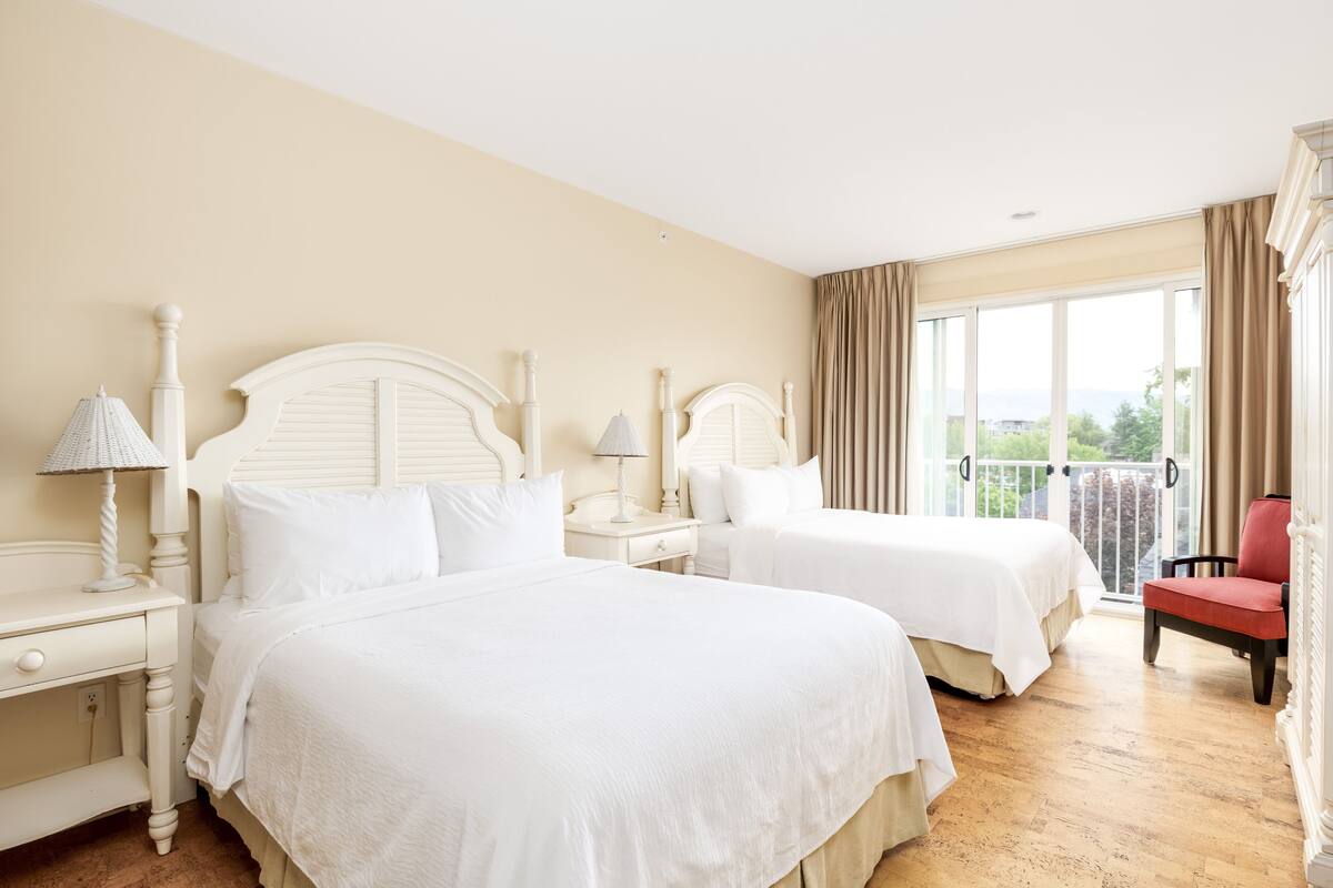 Ein Hotelzimmer mit zwei Betten, einem roten Sessel, einem Balkon mit Vorhängen und Blick auf Grünflächen.