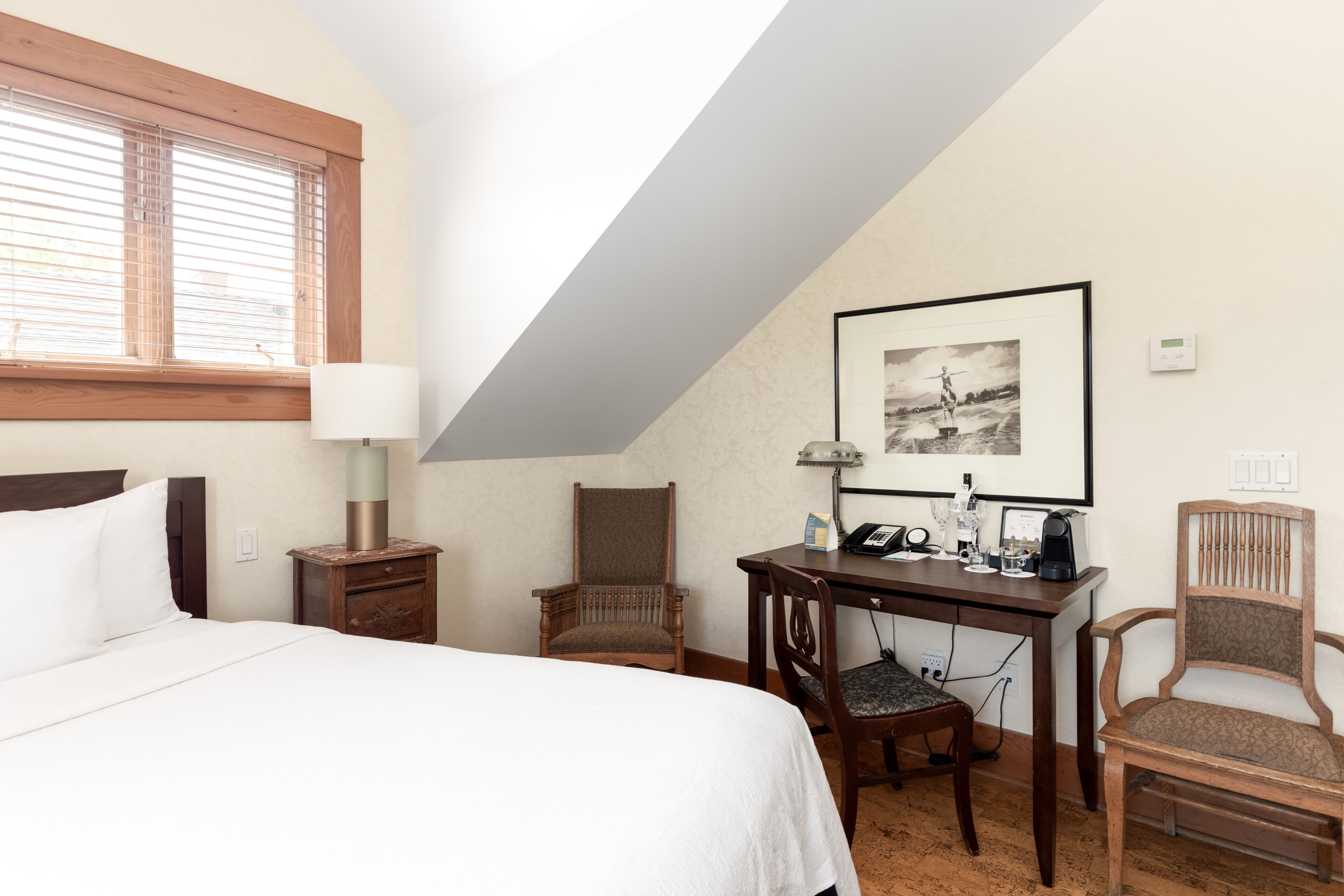 Hotelkamer met een bed, een bureau, twee stoelen, een lamp, een lijst met een foto aan de muur en een thermostaat.