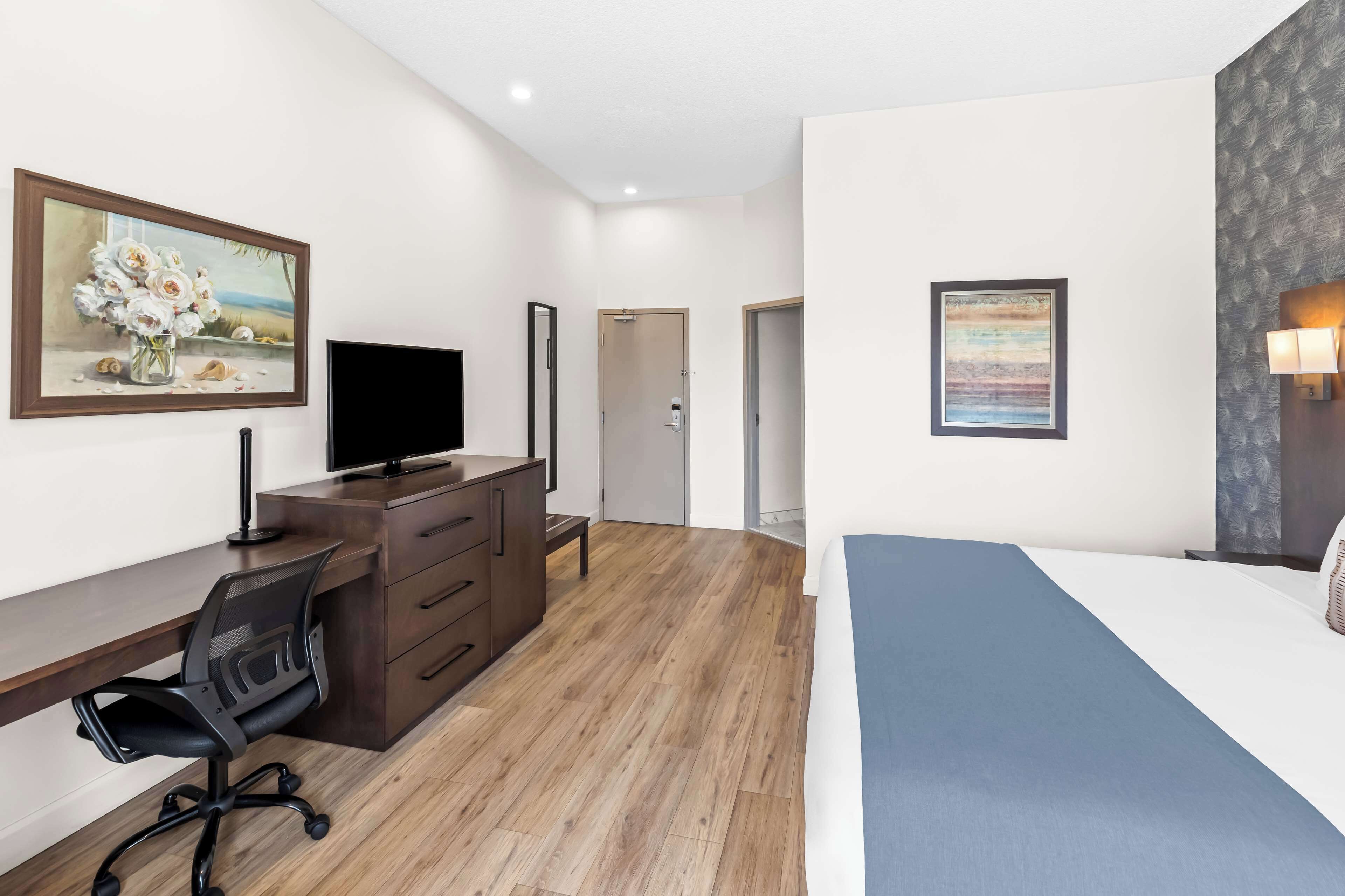 Una habitación de hotel moderna con suelo de madera, un escritorio con silla, una cama con colcha azul y dos cuadros enmarcados en las paredes.