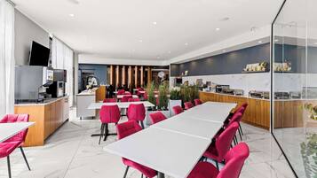 Un espace de restauration moderne dans un hôtel, avec une longue table, des chaises rouges, une cuisine en arrière-plan et une cloison en verre.