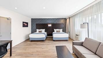 Una habitación de hotel moderna con dos camas, un sofá, un televisor y un amplio ventanal con cortinas.