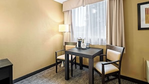 A hotel room with a dark wooden table, two beige chairs, a lamp, and a window with beige curtains.