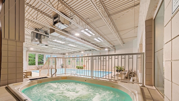 An indoor swimming pool with a hot tub, surrounded by a glass railing, and a view of an outdoor pool area.