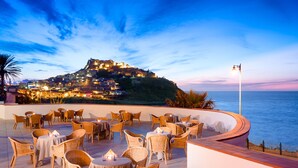 An outdoor dining area with wicker furniture and tables, overlooking a coastal town with buildings on a hill. - LH Pedraladda Resort (Castelsardo)