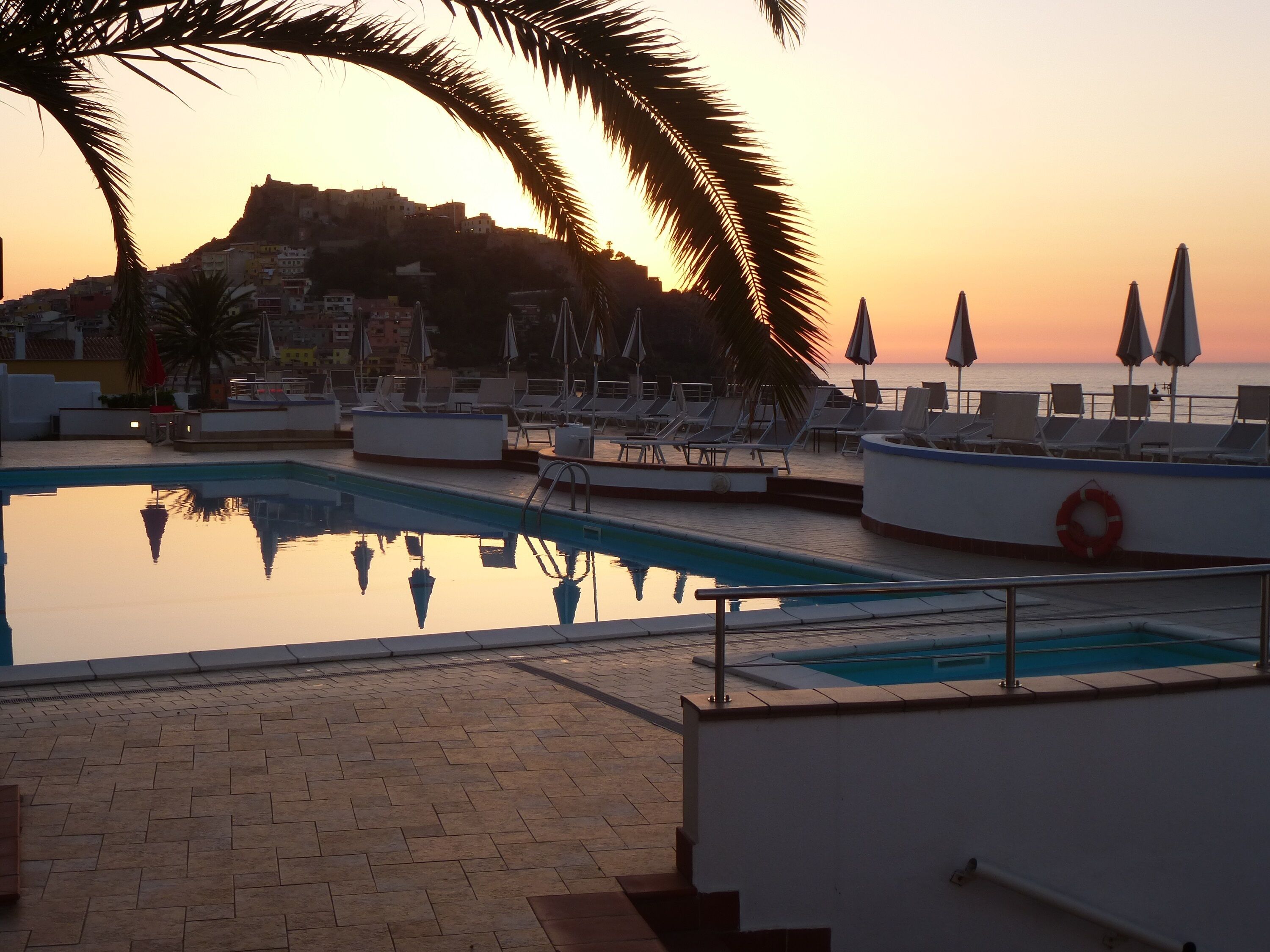 A swimming pool with lounge chairs and umbrellas, a lifebuoy, and a view of the sea at sunset.