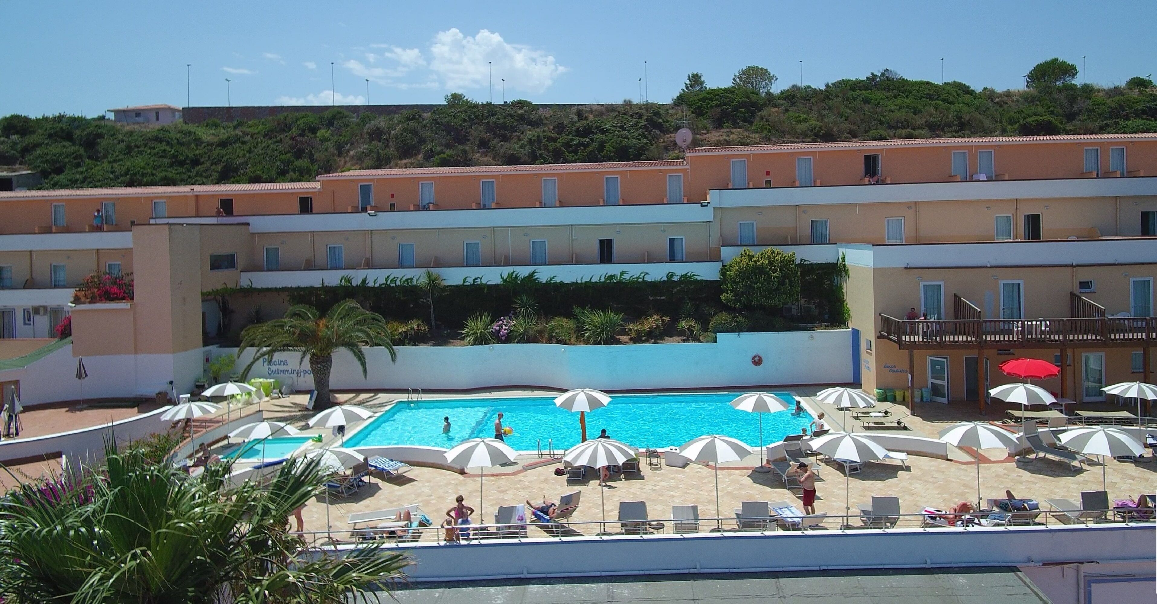 Ett hotell med en swimmingpool omgiven av solstolar och parasoller, en strand och utsikt över en sluttning.