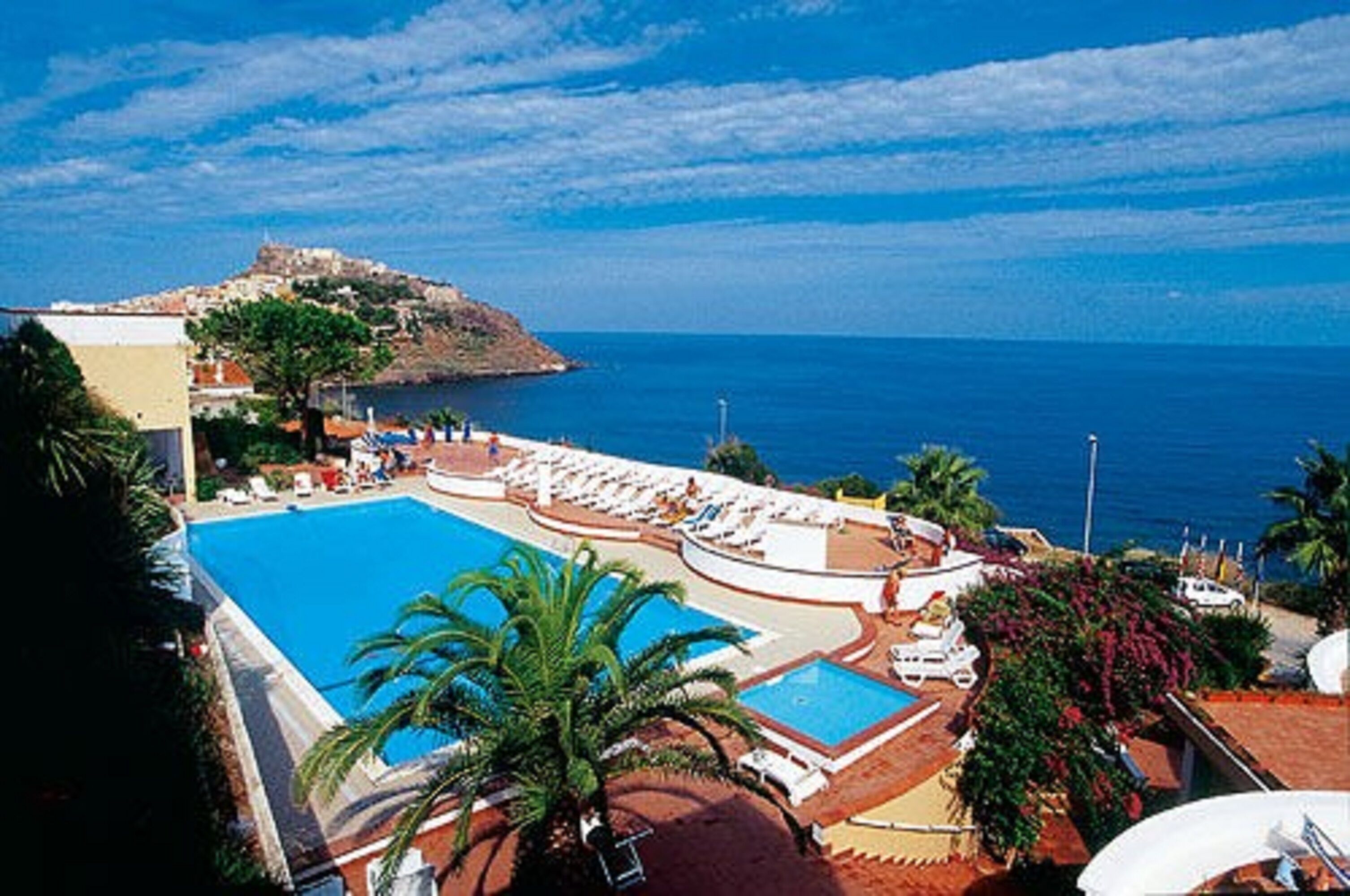 Un resort con una piscina grande, tumbonas y vistas al mar y a una colina.