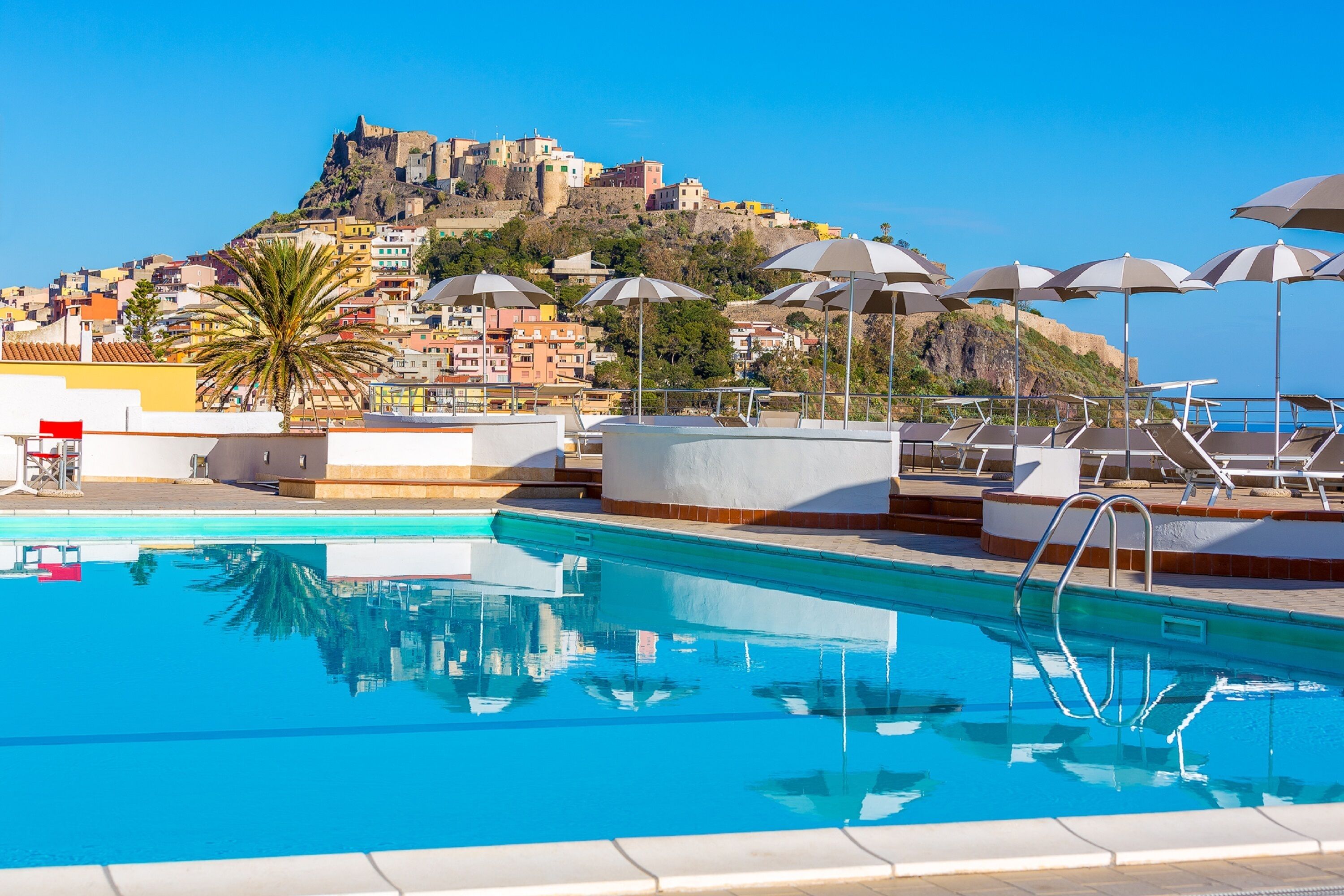 A swimming pool with lounge chairs and umbrellas, a clear blue sky, and a hill with buildings in the background.