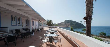 An outdoor terrace with tables and chairs, overlooking a coastal view with a palm tree and a hill in the distance.