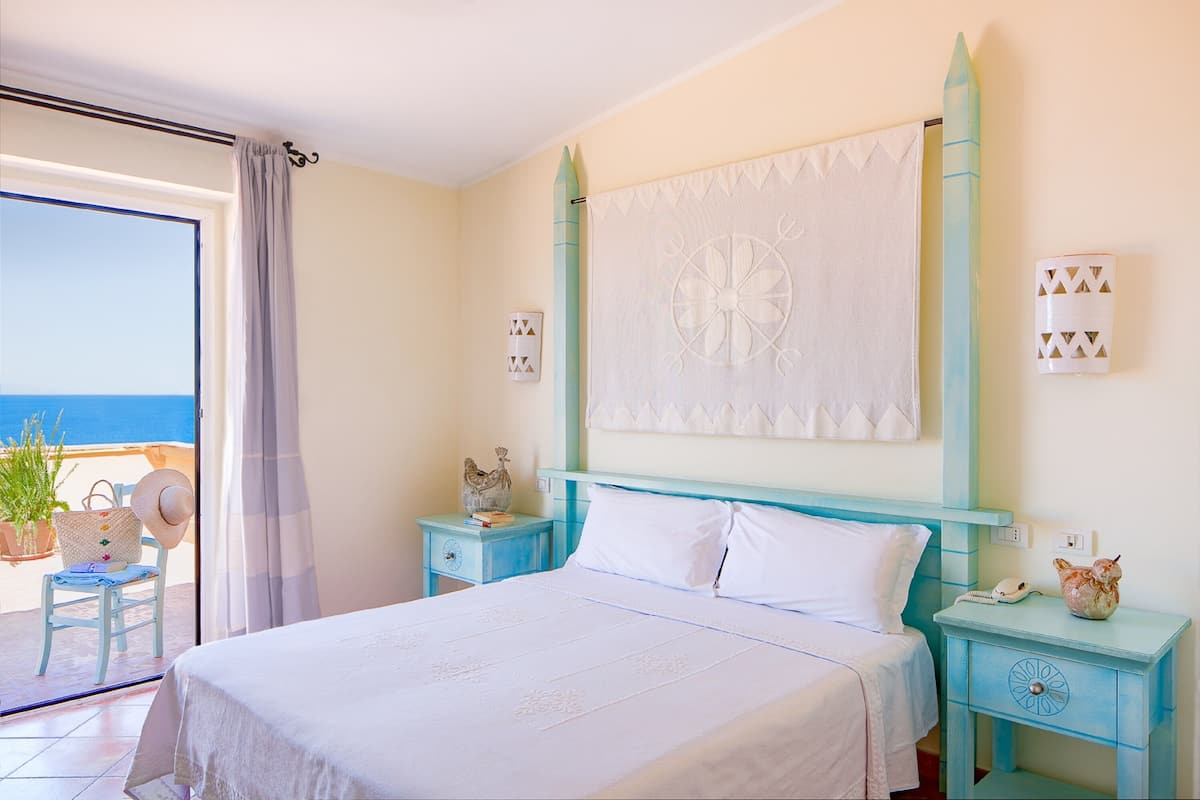 A bedroom with a bed, a nightstand, a chair, and a view of the sea.