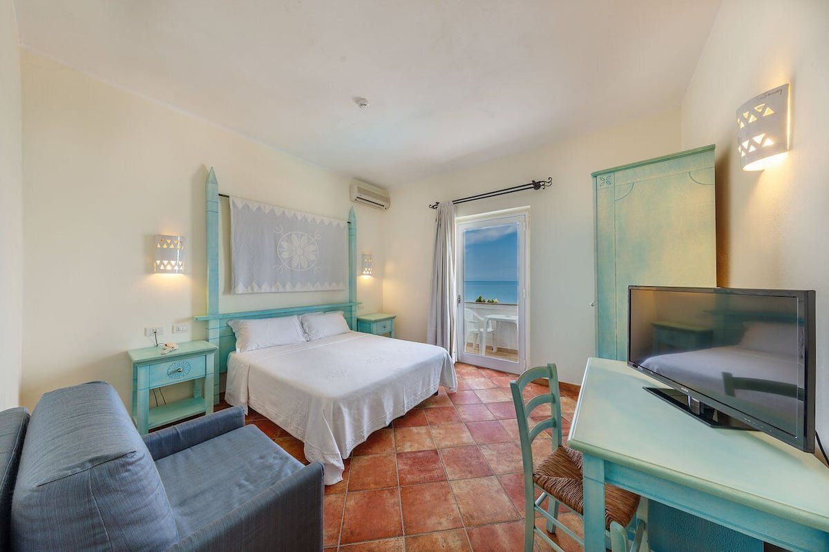 A bedroom with a bed, a TV, a desk, and a chair. There is a window with a view of the sea.