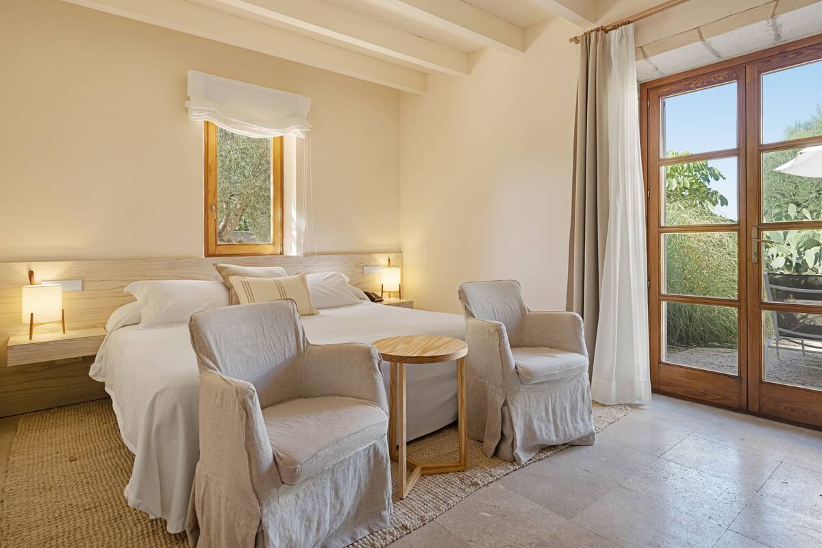 Une chambre à coucher avec un grand lit, deux tables de chevet avec des lampes, une petite table ronde, deux fauteuils, une grande fenêtre avec des rideaux et une vue sur la verdure à l’extérieur.