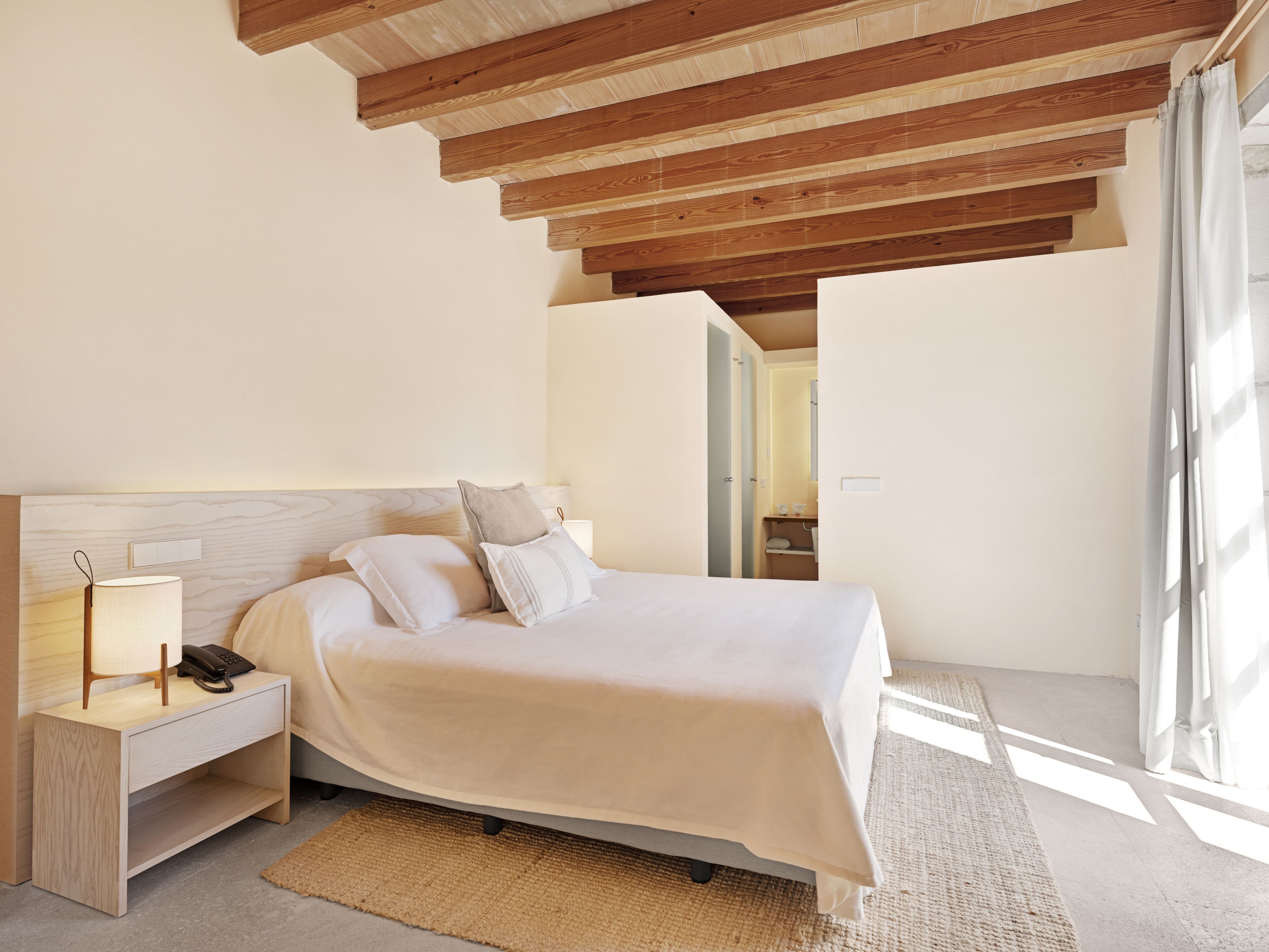 Un dormitorio con techo de madera, una cama con ropa de cama blanca, una mesita de noche con lámpara y vistas al baño.