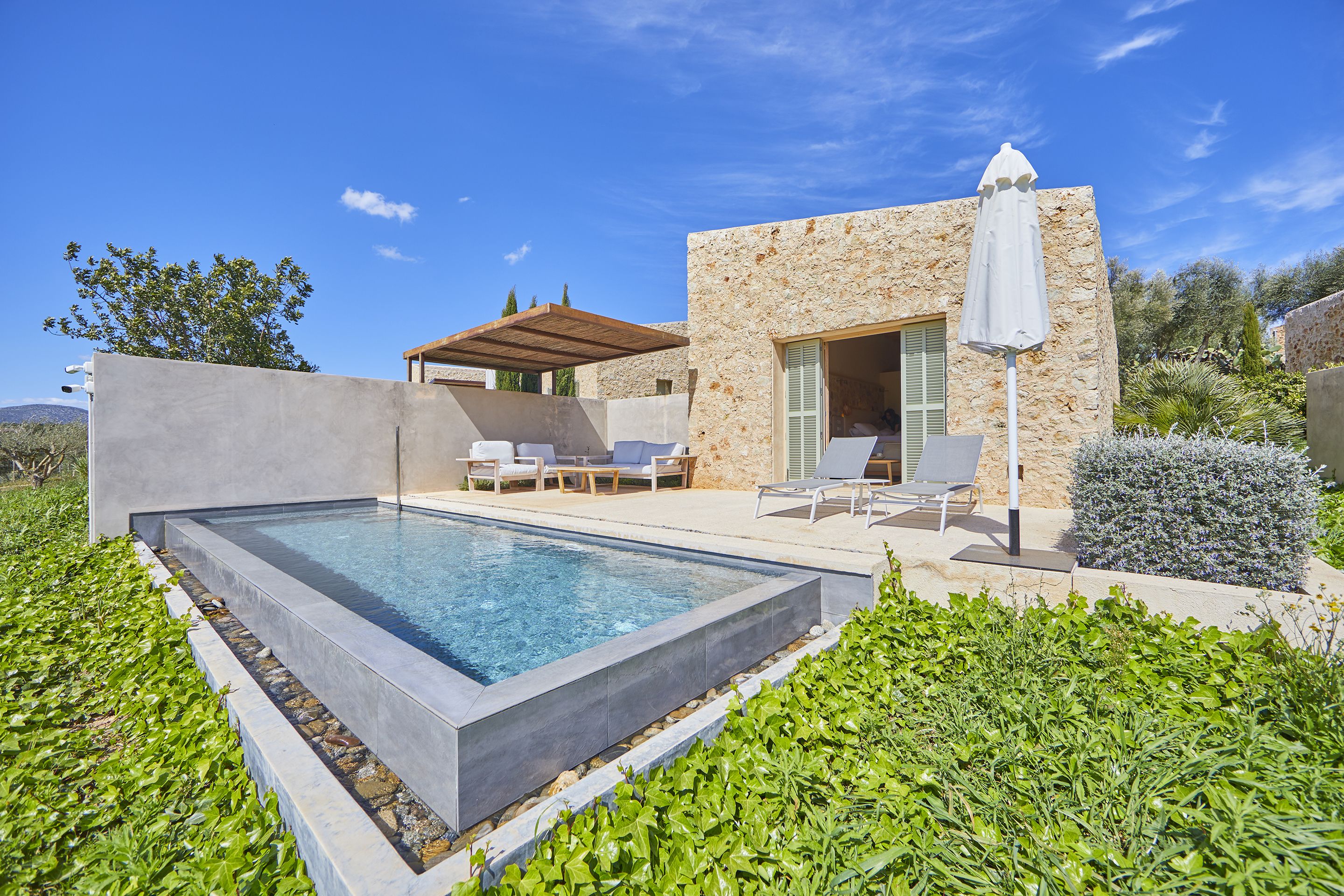 A modern pool area with a stone building, outdoor seating, and a clear blue sky.