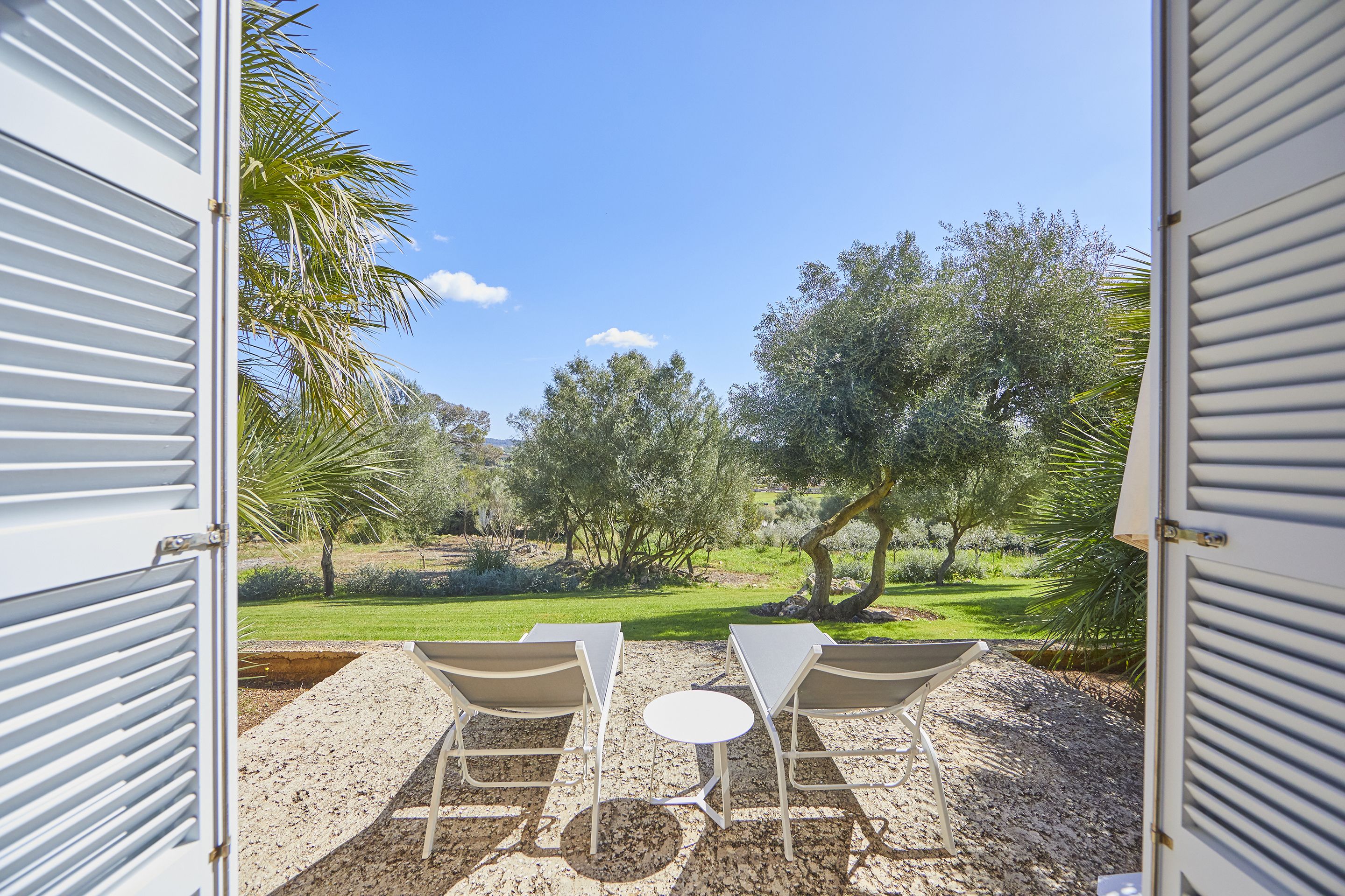 An open patio with two white tables and chairs, a gravel floor, and a view of a green landscape with trees and a clear sky.