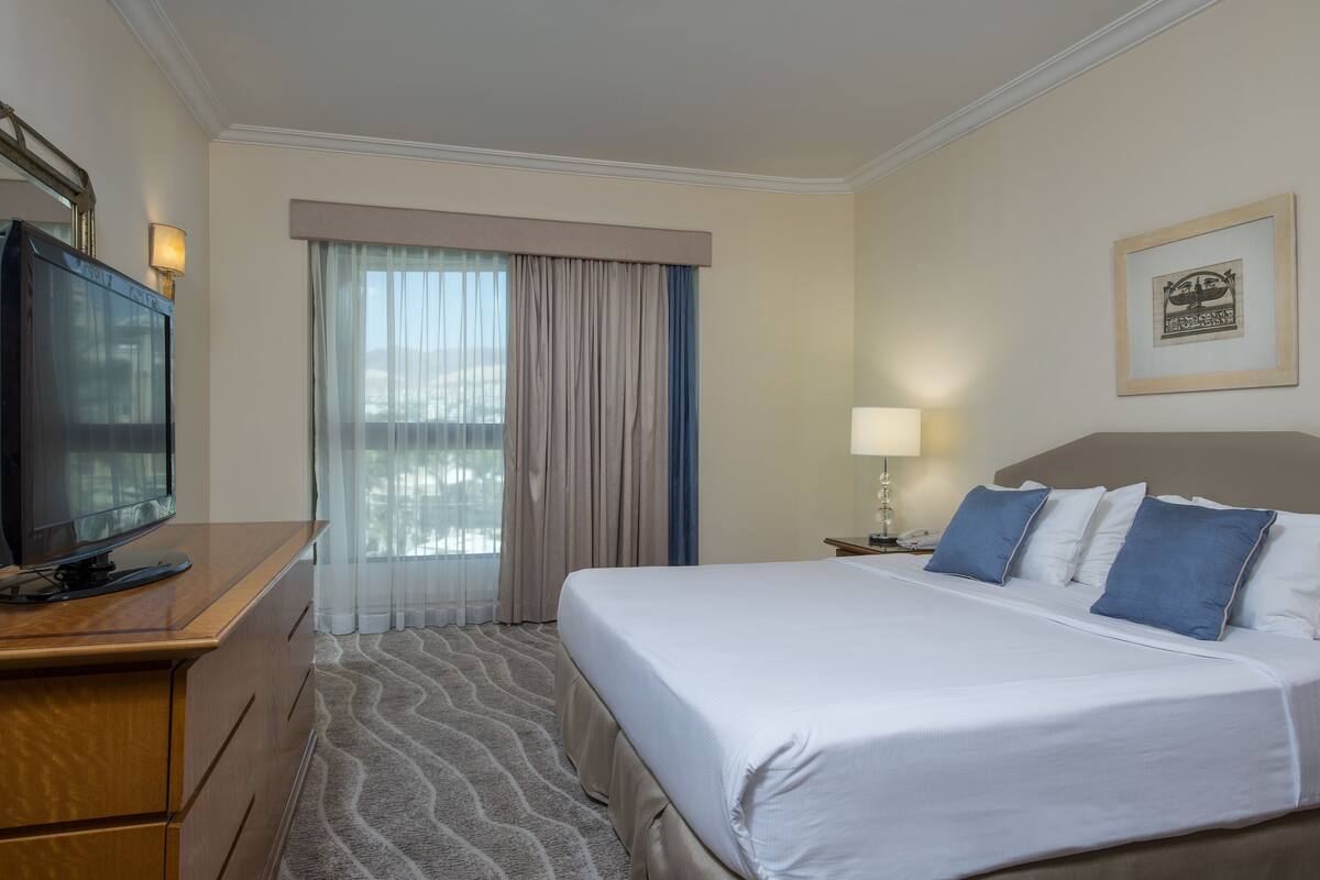 Una habitación de hotel con cama, televisión, escritorio, lámpara y ventana con cortinas.