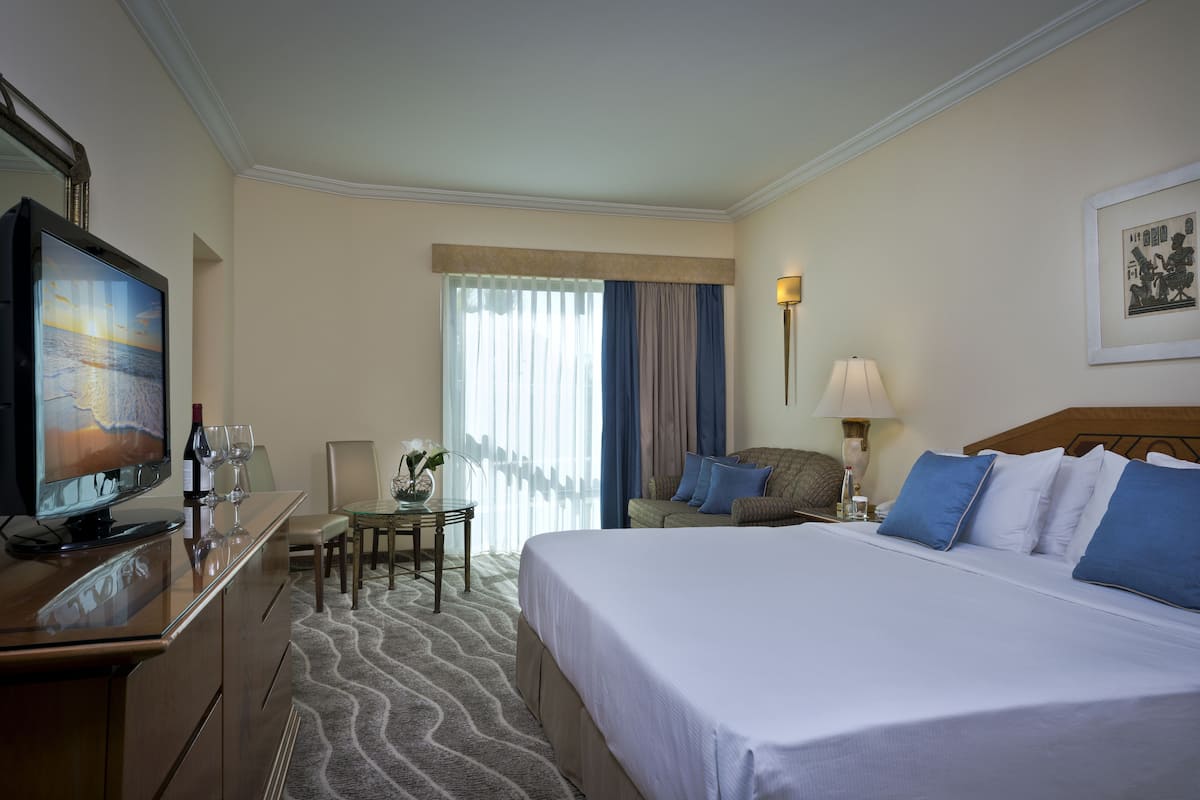 Una habitación de hotel con una cama, un escritorio con televisor, una silla, una mesa con un jarrón y una ventana con cortinas.