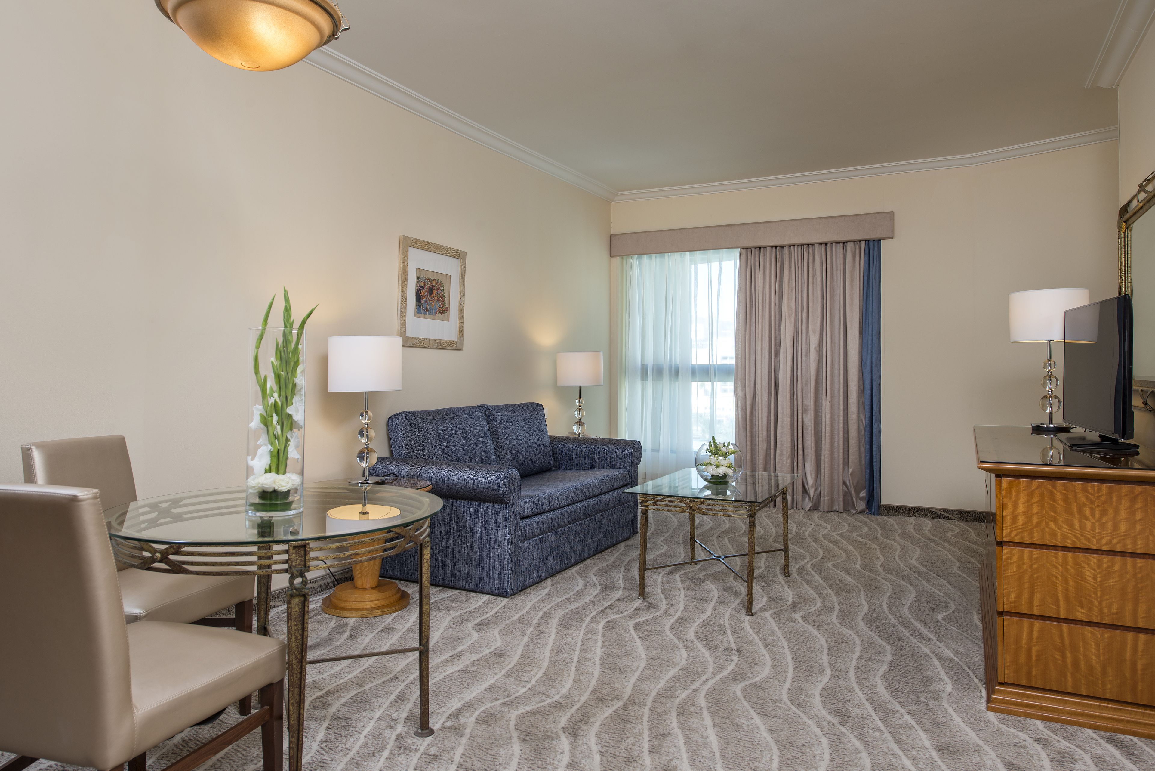Ein Hotelzimmer mit einem runden Glastisch, einem blauen Sofa, einem kleinen Beistelltisch, einer Holzkommode und einem großen Fenster mit Vorhängen.
