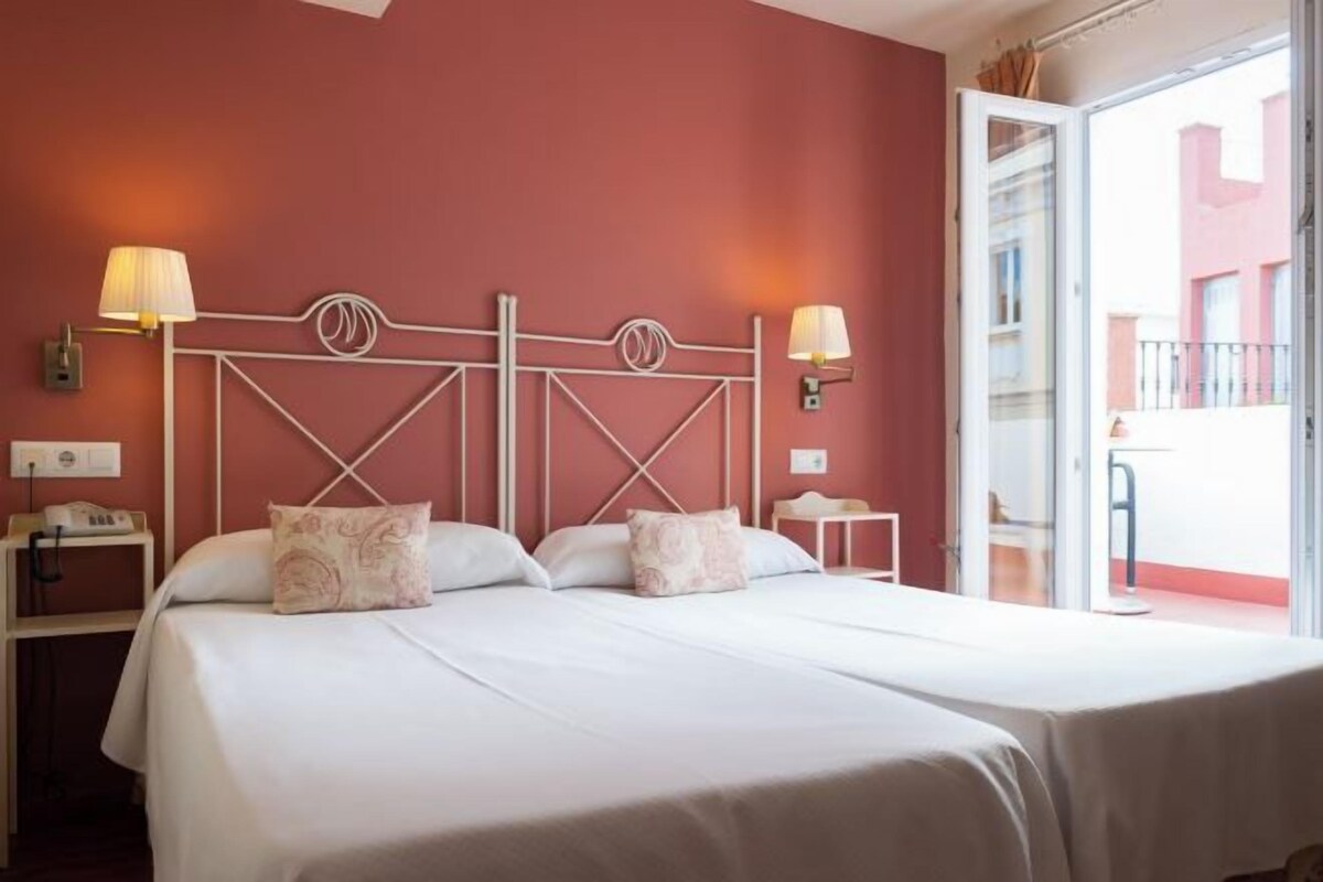 Een hotelkamer met een bed, nachtlampjes, een nachtkastje en uitzicht door een geopende deur.
