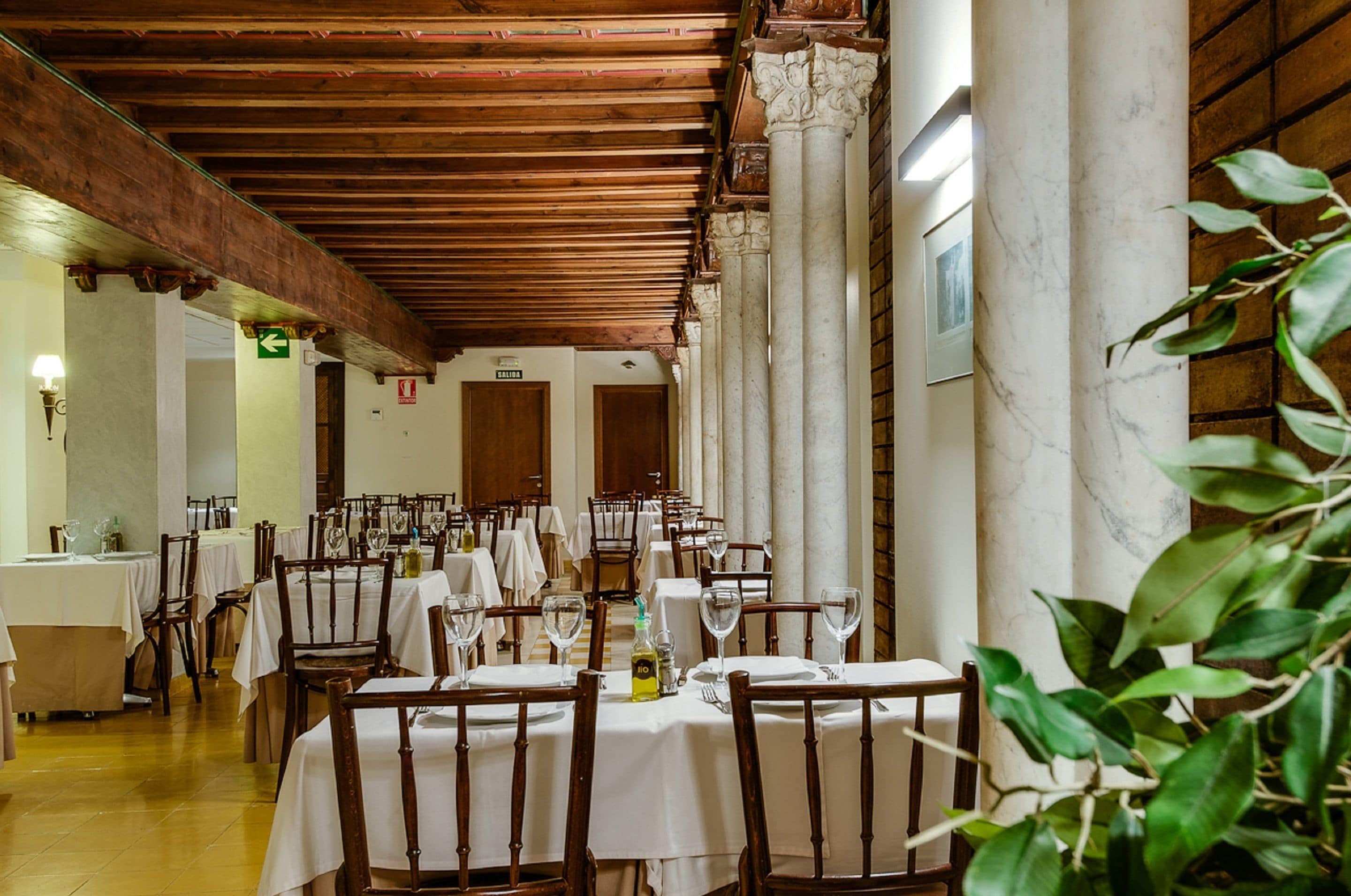 A restaurant with multiple tables and chairs, wooden beams on the ceiling, marble columns, and a green plant in the foreground.
