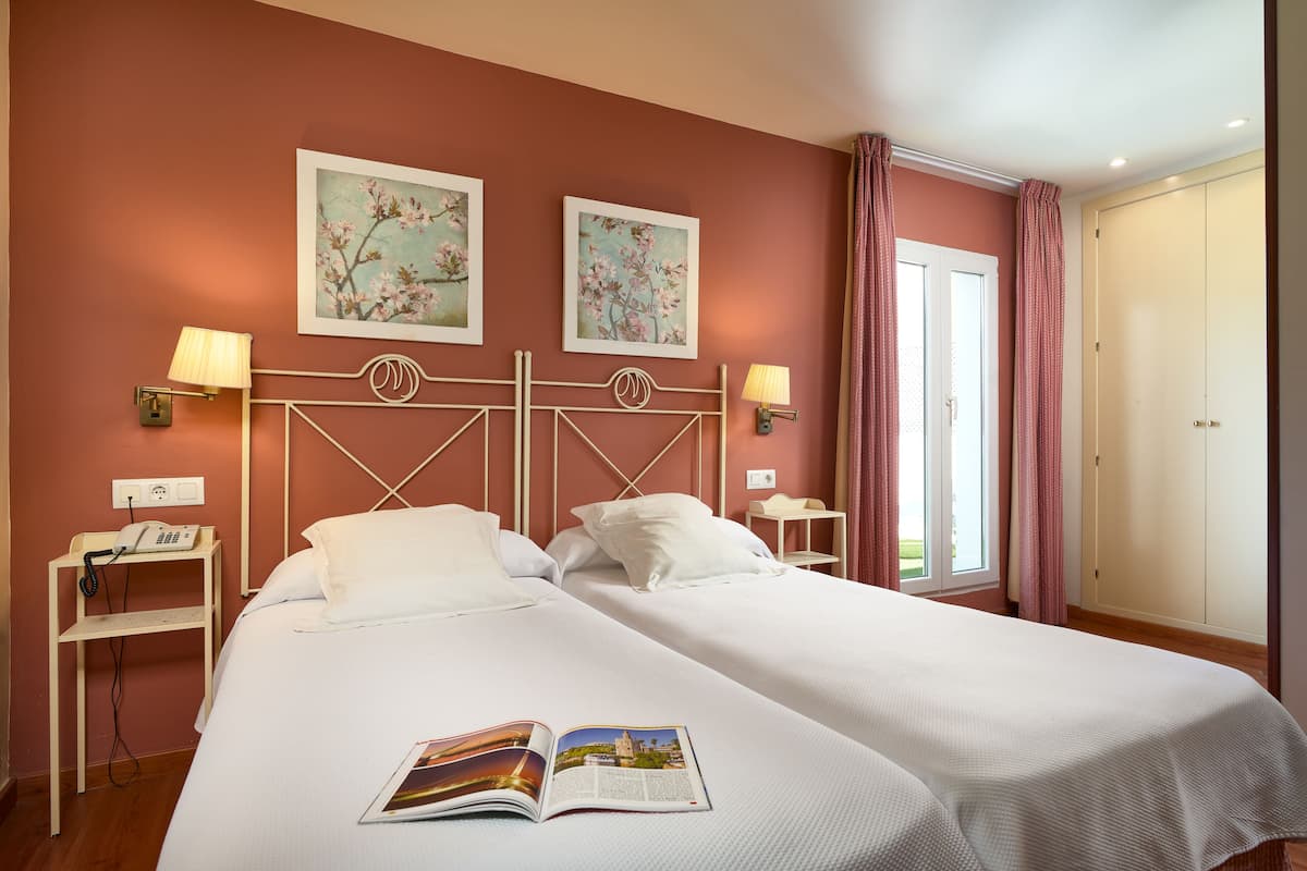 Un lit double avec du linge de lit blanc, deux lampes fixées au mur, une table de chevet avec un téléphone et deux tableaux représentant des motifs floraux, encadrés.