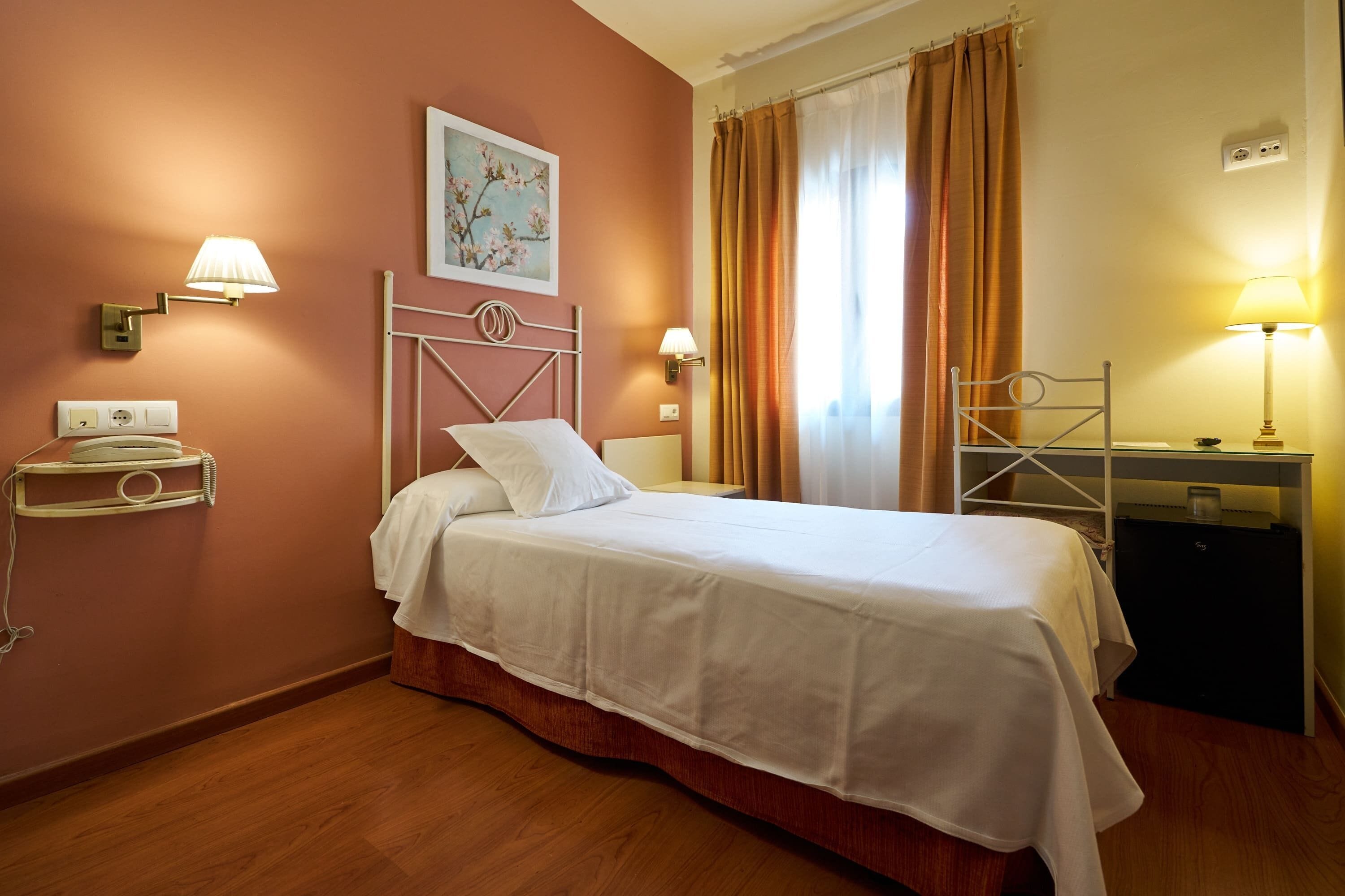 Een hotelkamer met een enkel bed, nachtkastje, lamp, wandplank en schilderij.