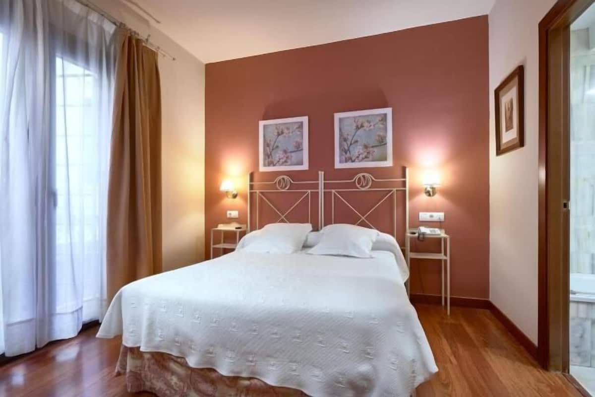 Une chambre d’hôtel avec un lit, deux lampes fixées au mur, une table de chevet, une fenêtre avec des rideaux et deux tableaux encadrés accrochés au mur.