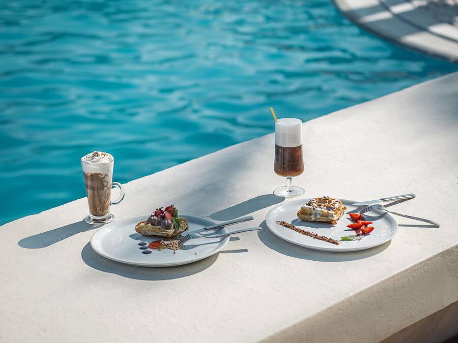 A dining table by a pool with two plates of food and two drinks.