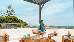 Een terras met een houten tafel, een witte bank en rieten stoelen, met uitzicht op een schilderachtig zeegezicht.