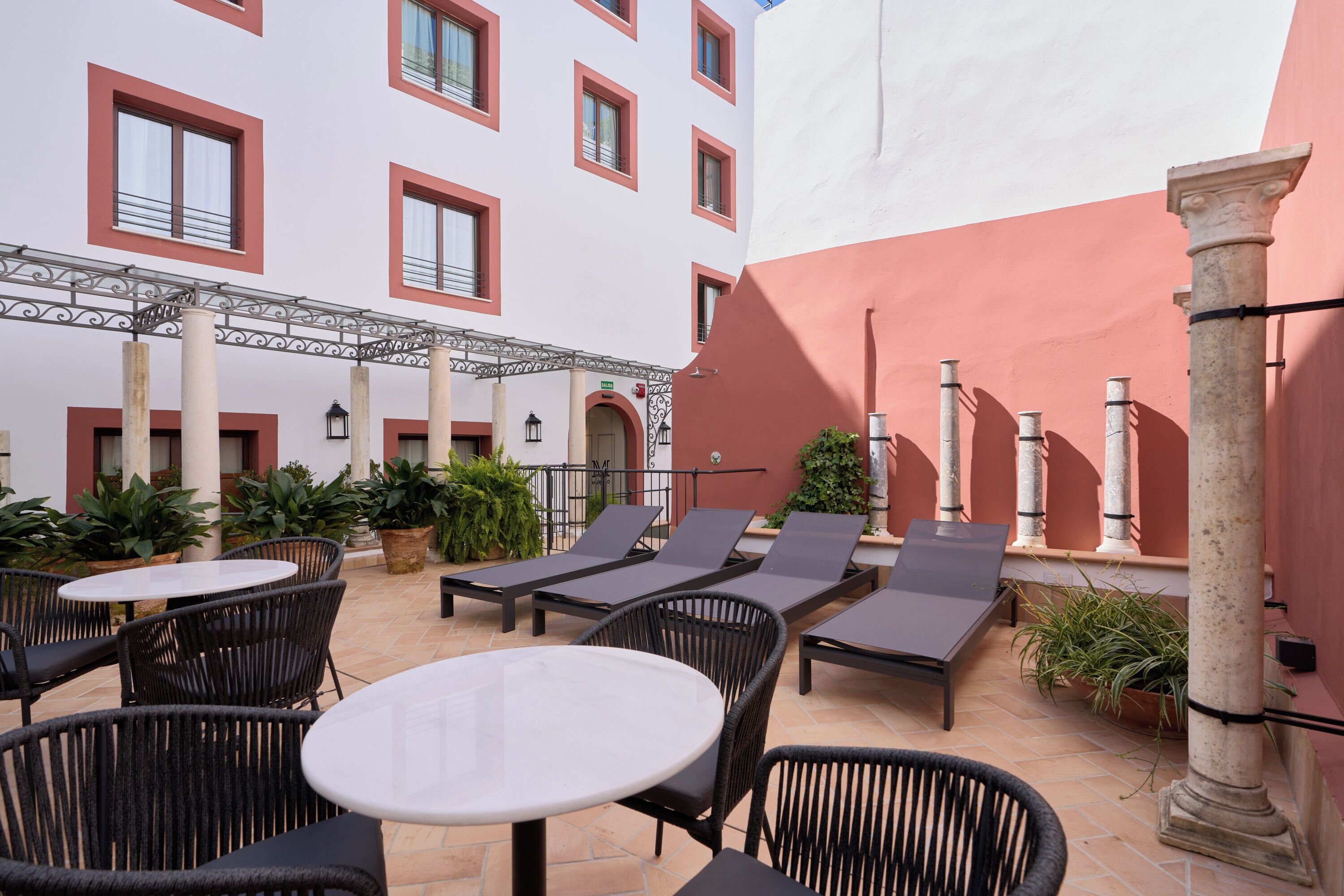 En udendørs patio med loungesenge og borde, omgivet af plantede krukker og arkitektoniske søjler.