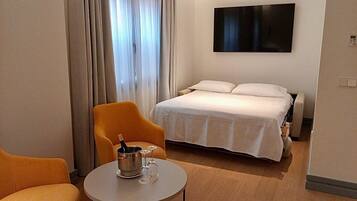 A hotel room with a bed, a chair, a small table with a bottle and glasses, and a TV on the wall.
