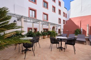 An outdoor patio with tables and chairs, surrounded by potted plants and a building with red-framed windows. - Hotel Murillo Reinoso (Seville)