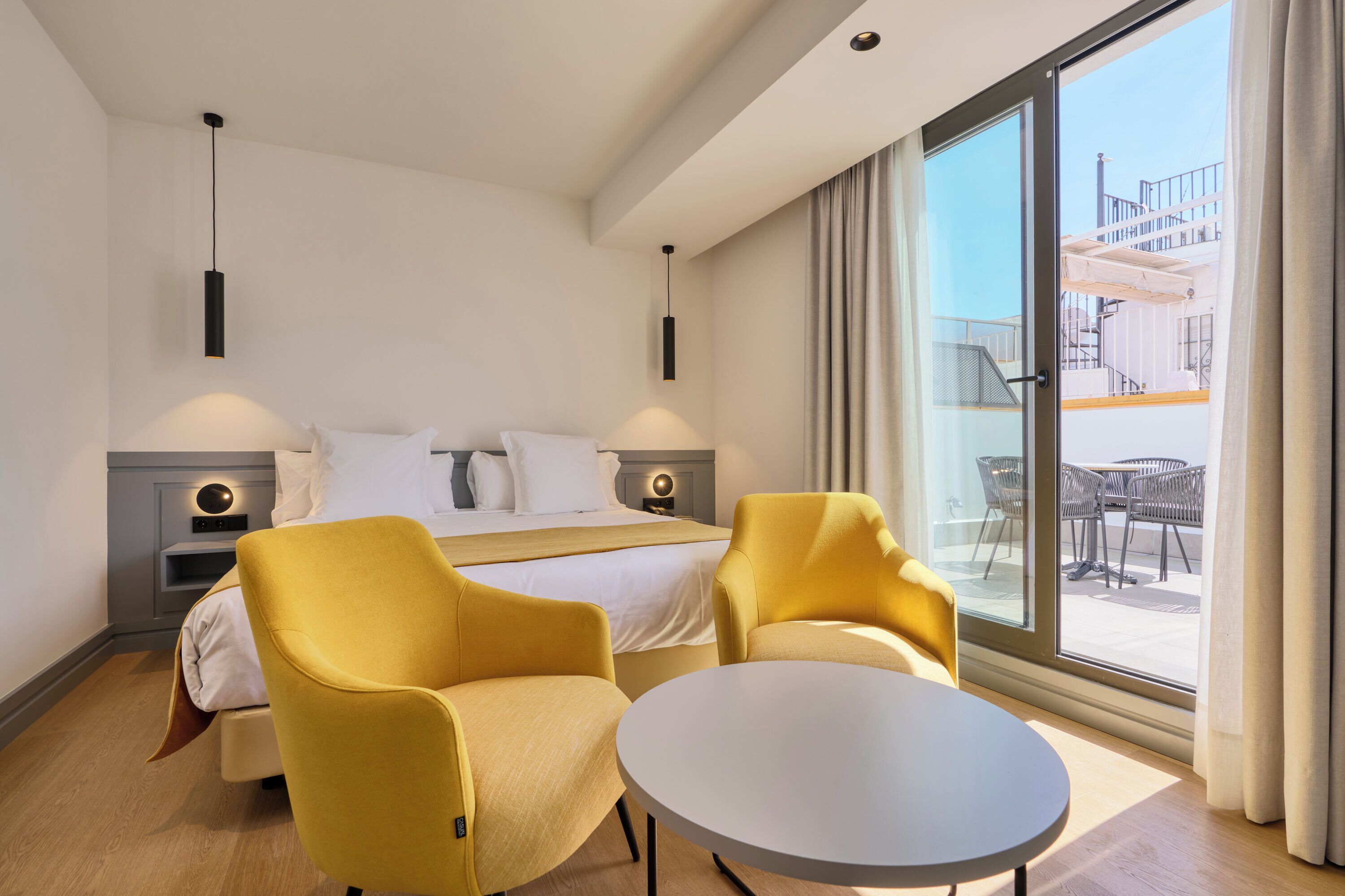 Habitación de hotel con cama, dos sillones amarillos, una mesa redonda y un ventanal con vistas a muebles de exterior.