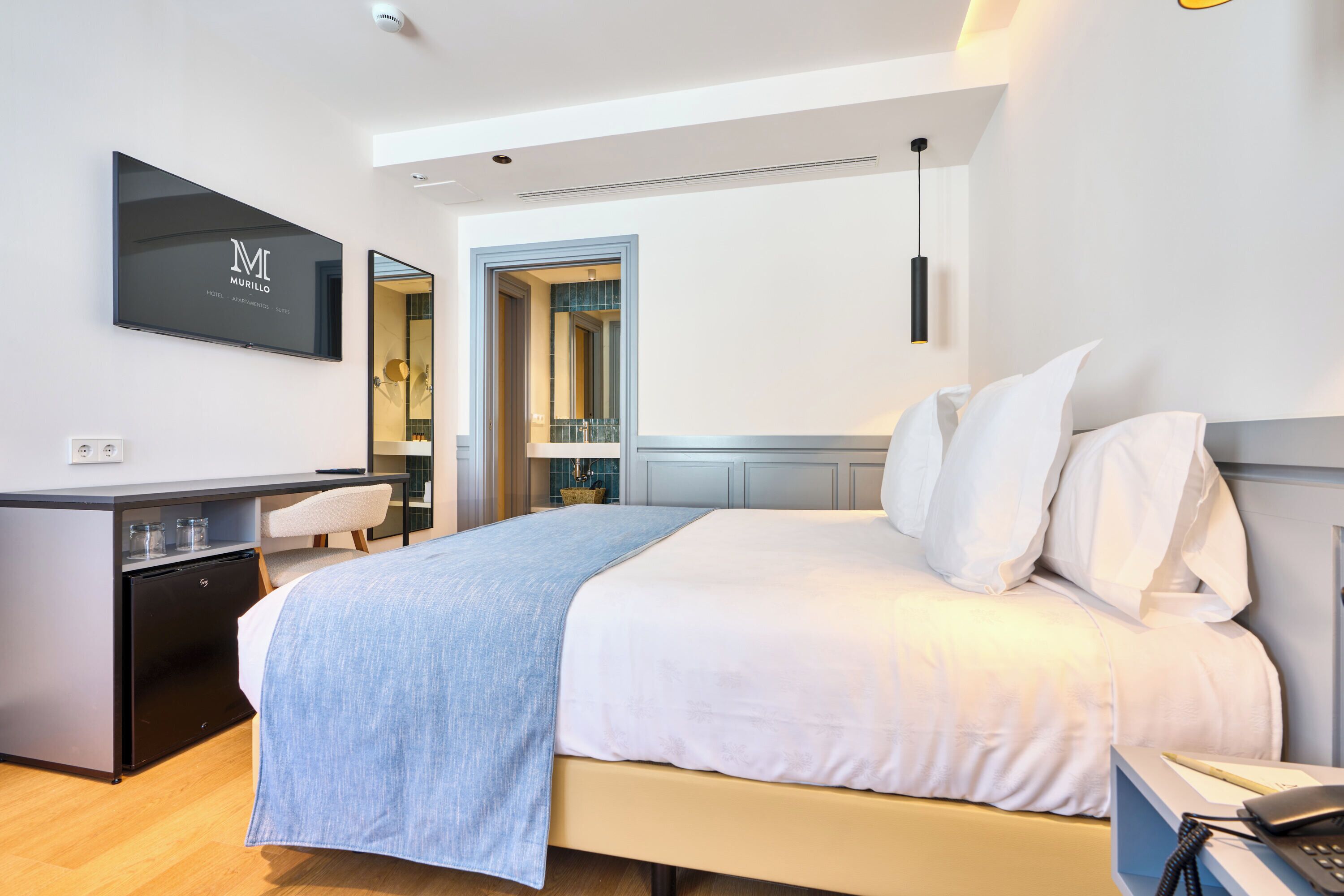 Habitación de hotel con cama, escritorio, silla, televisor y baño visible a través de una puerta abierta.