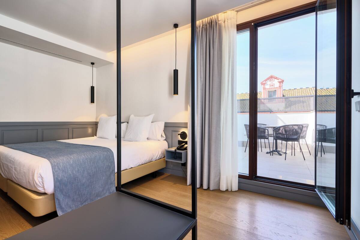 Una habitación de hotel moderna con cama, mesita de noche y vistas al área de estar exterior a través de un ventanal.