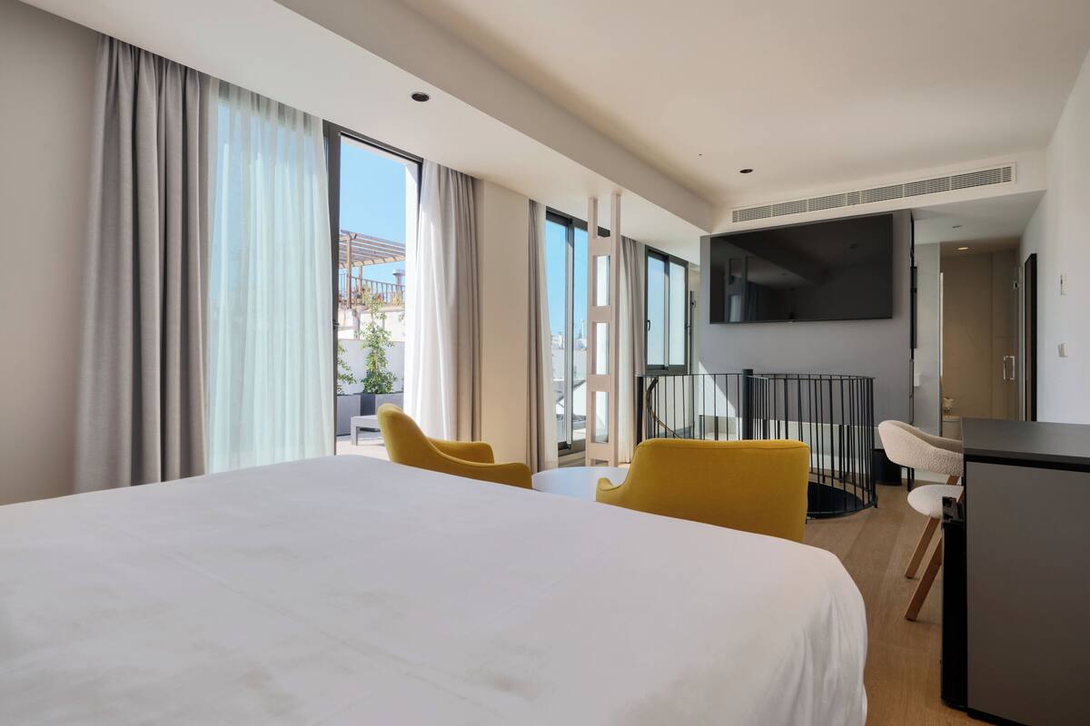 Habitación de hotel con una cama grande, televisión, escritorio y balcón con sillas amarillas.