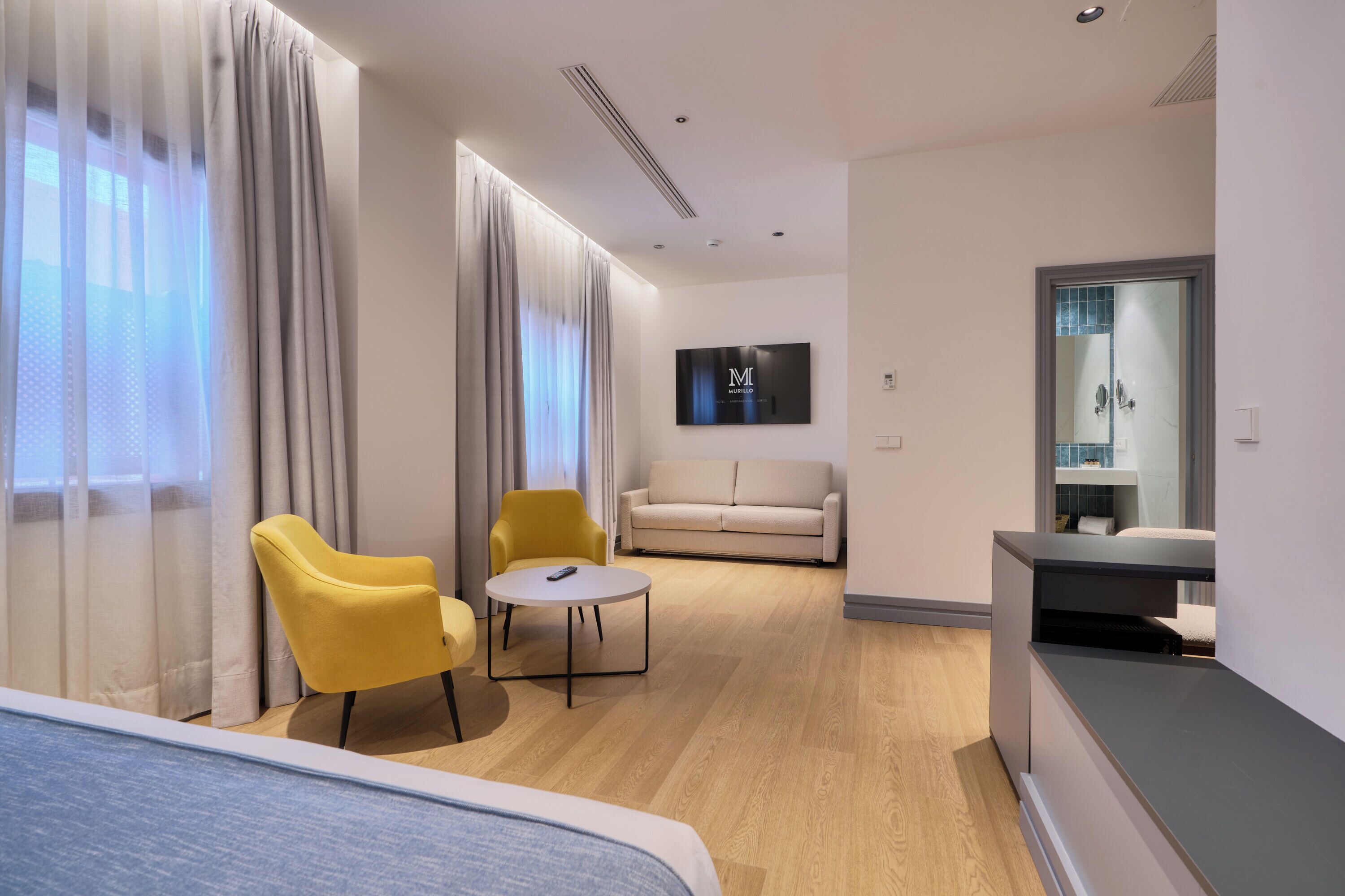 Una habitación de hotel moderna con una cama, un sillón amarillo, una pequeña mesa redonda, un sofá y un televisor montado en la pared.
