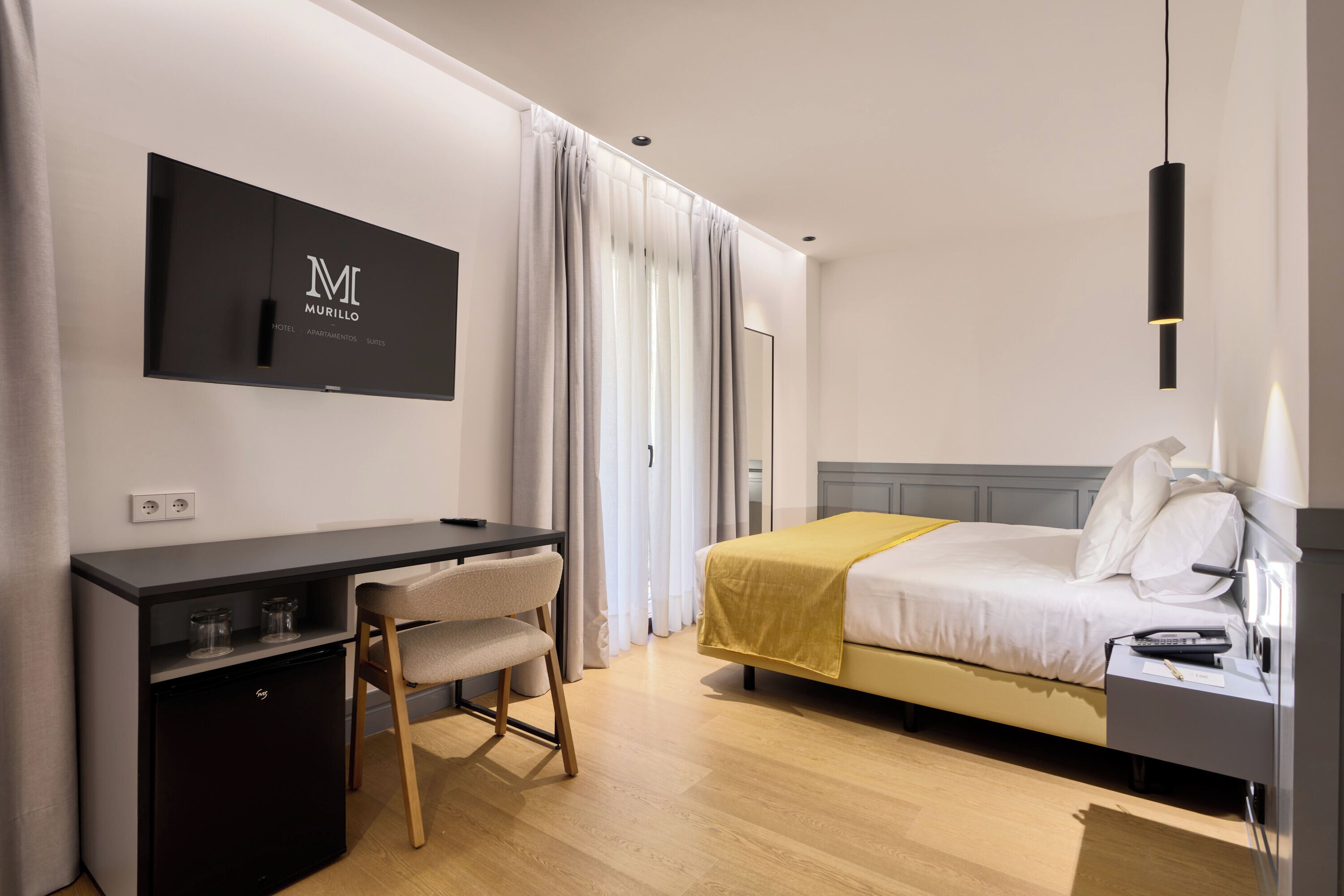 A hotel room with a bed, a desk with a chair, a TV mounted on the wall, and a wardrobe.