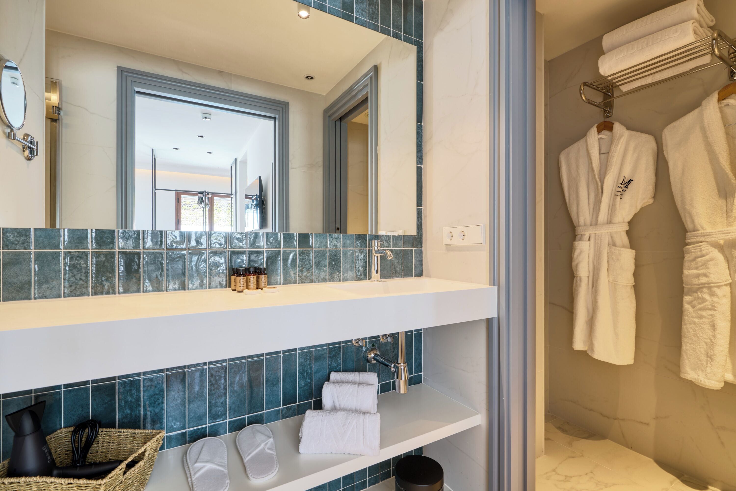 Un baño con un espejo grande, un lavamanos y una zona de ducha. Hay toallas y una cesta con artículos de aseo en la encimera.