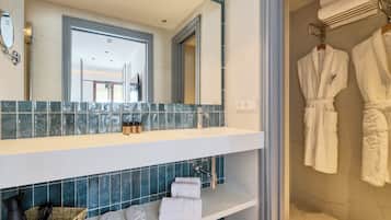 Un baño con un espejo grande, un lavamanos y una zona de ducha. Hay toallas y una cesta con artículos de aseo en la encimera.