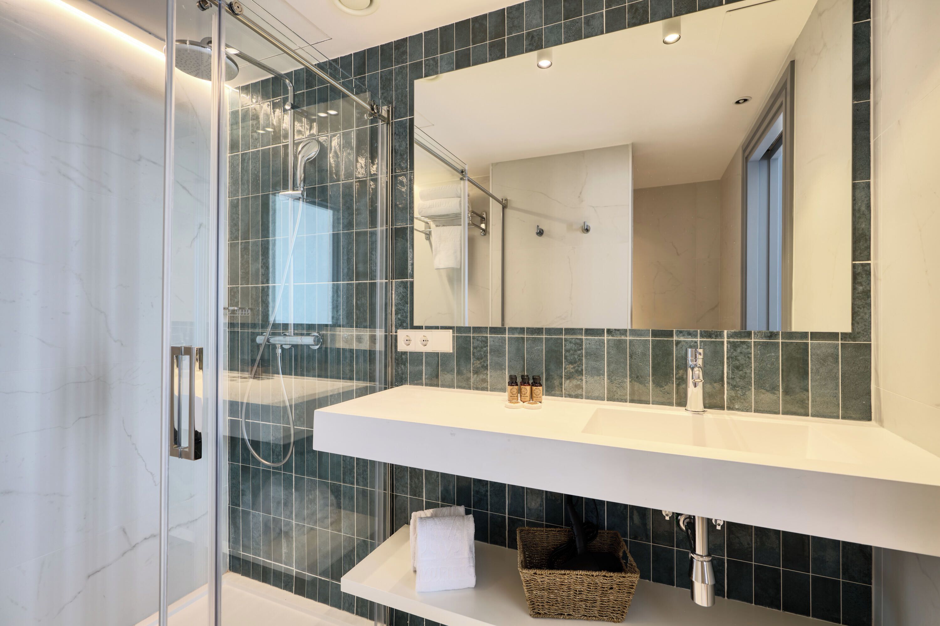 Un baño moderno con ducha envuelta en vidrio, tocador blanco con espejo y paredes cubiertas de azulejos verde oscuro.